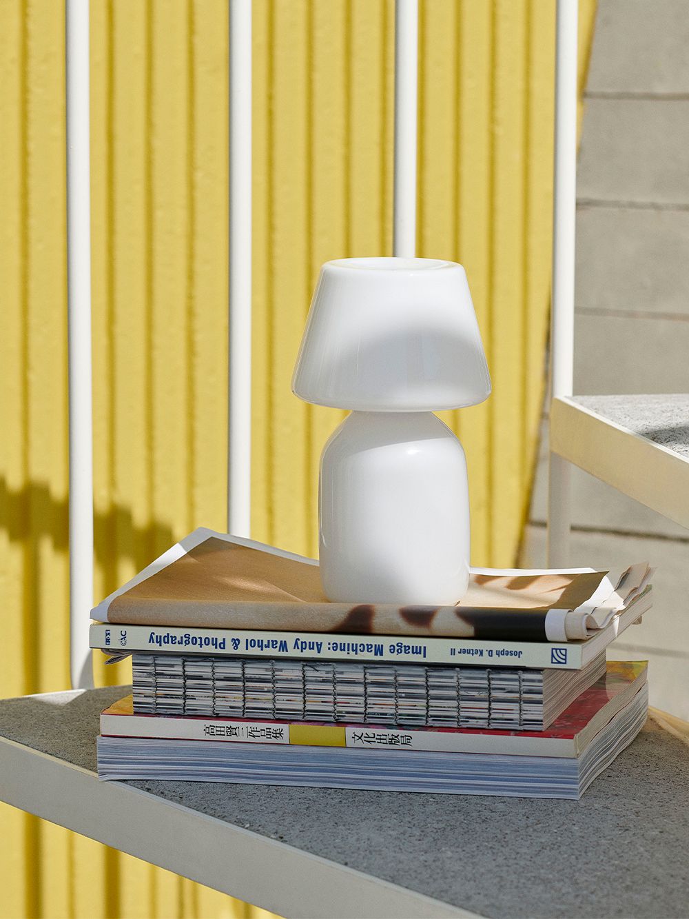 HAY’s opal white Apollo table lamp stands on top of magazines outdoors in the sunshine.