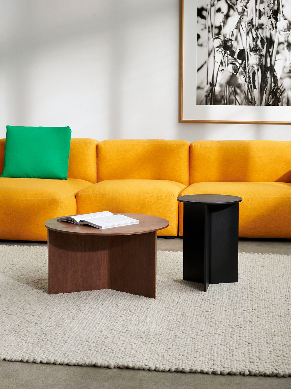 There are two Slit Wood tables in front of an orange sofa, a lower one in dark wood and a higher one in black. There is an open book on the wood-colored table.