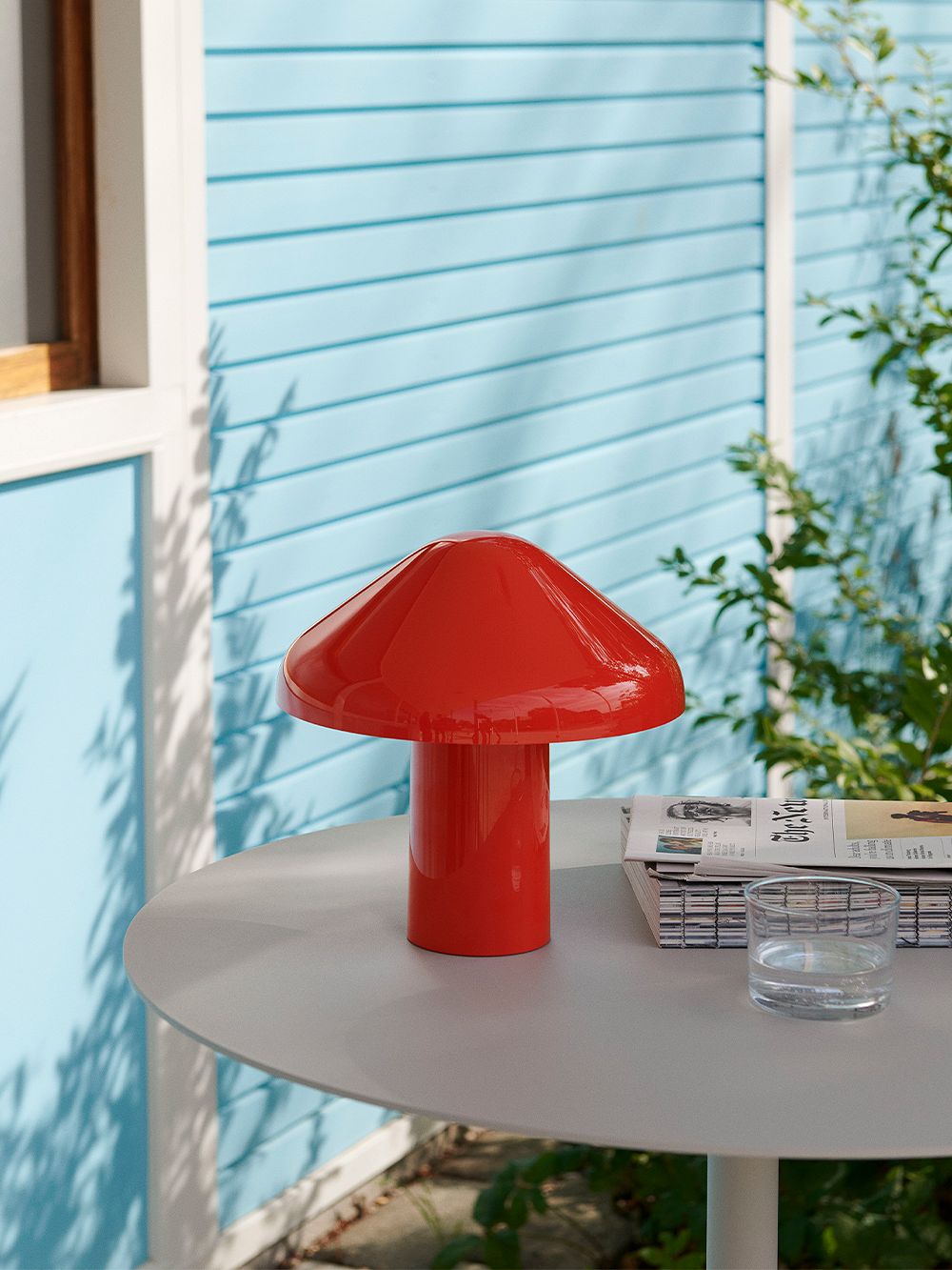 The red Pao Portable lamp on a table outdoors in the sunshine, with a light blue wall in the background.