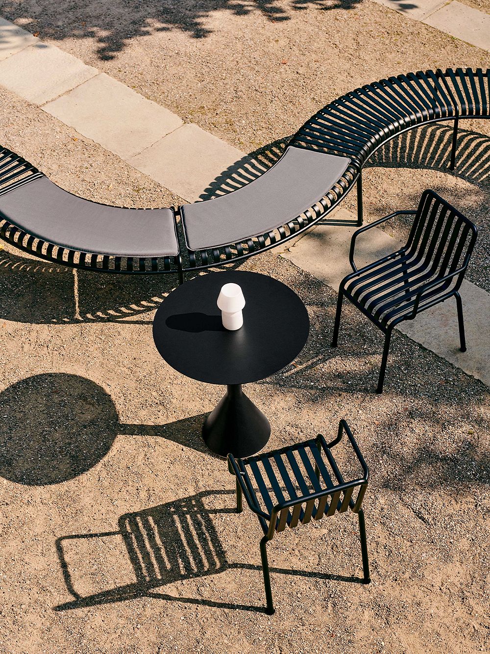 The table, chairs and benches in HAY's Palissade series make up a seating area on a sandy yard.