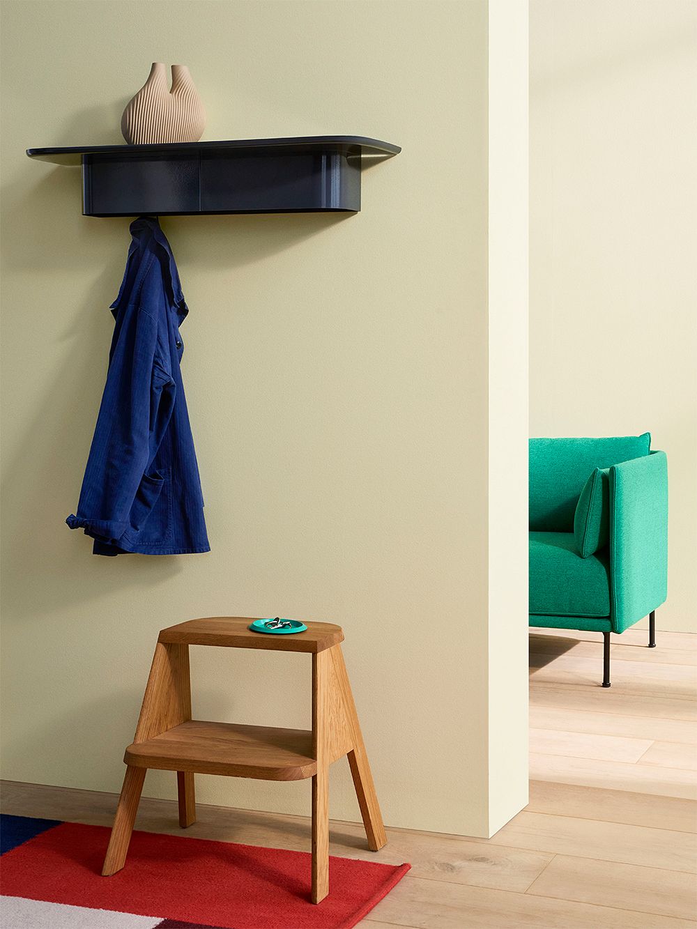 HAY's Korpus shelf on the hallway wall. There is a vase on top of the shelf, and a coat hangs on its hooks. Below is a wooden stepladder, and in the background is another room with a green sofa.