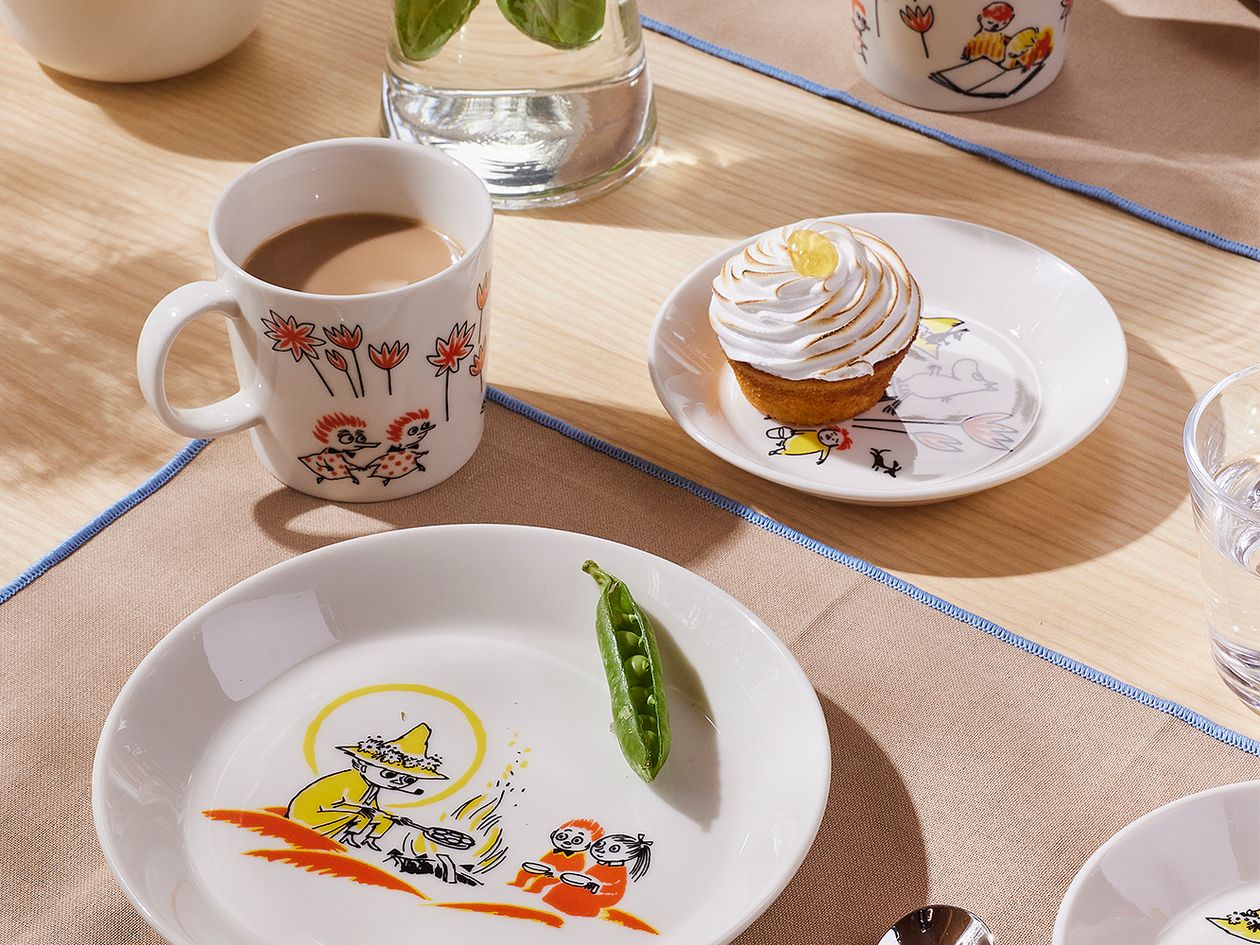 Table setting with Moomin by Arabia x Red Cross tableware. Coffee in a mug and a muffin on a plate.