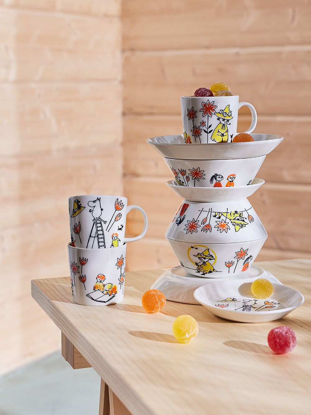 A quirky pile of Moomin by Arabia x Red Cross tableware on a wooden table: mugs, plates, bowls.