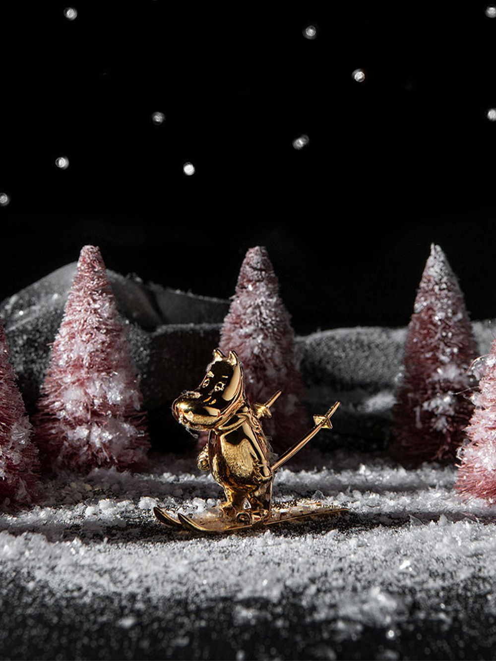 A gilded Moomintroll figurine with skis and sticks in a dark wintery lanscape with trees and hills in the background.