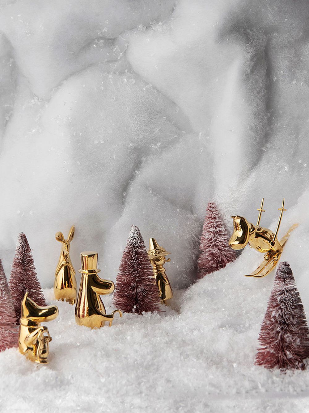 A gilded Moomintroll figurine skiing down a snowy hill with the rest of the Moomin family watching from between trees.