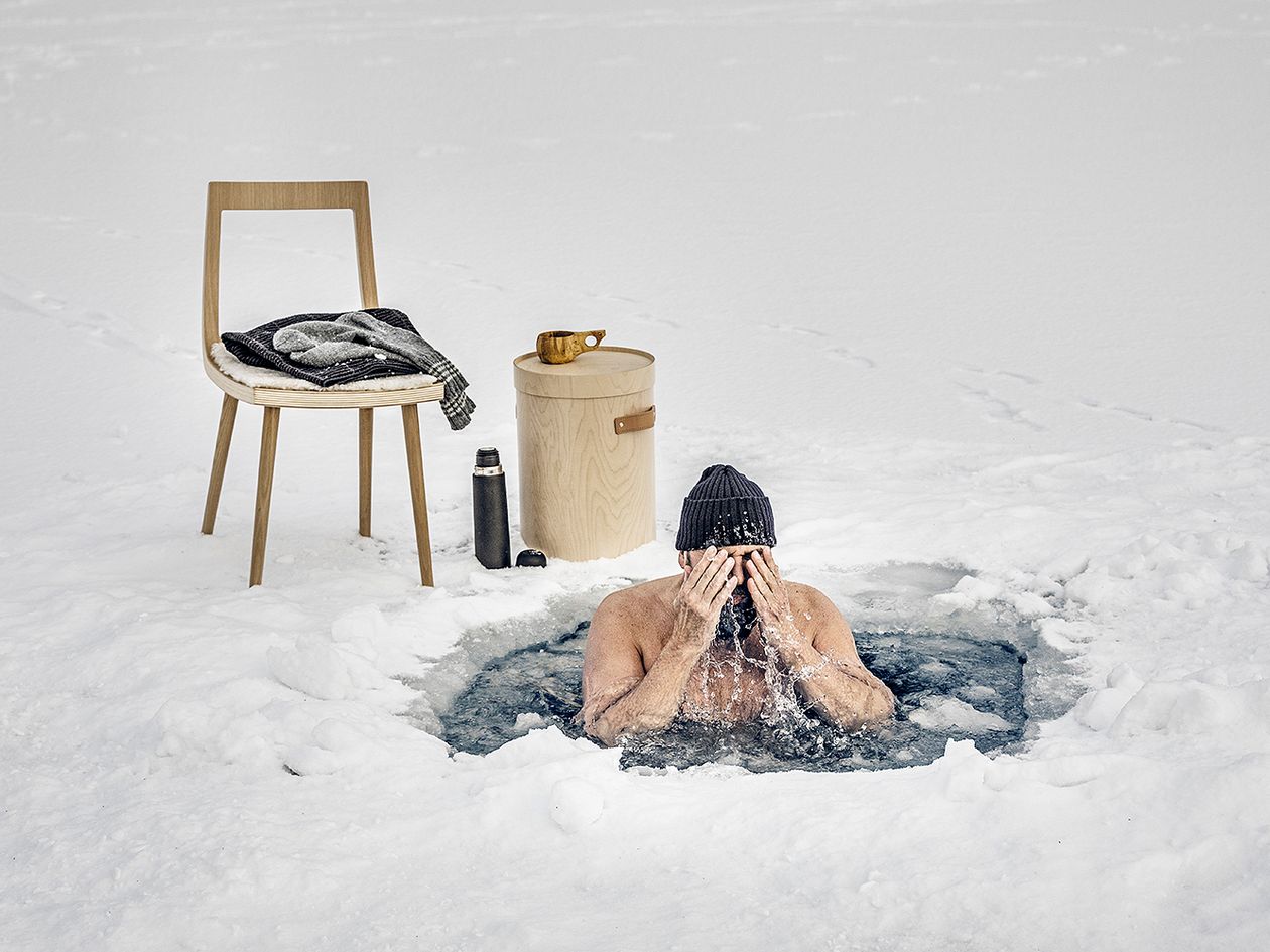 Designer Tapio Anttila in an ice hole wiping water off his face. In the background is his Viiva chair with a pile of clothes on it.