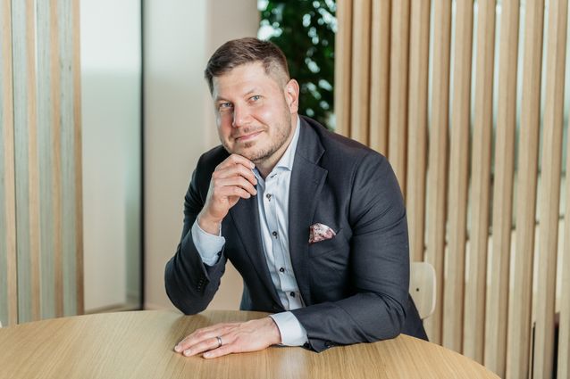 Kuvassa hymyilevä mies tummassa puvussa istuu ja nojaa pöytään.