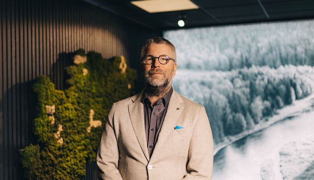 Kuvassa hymyilevä mies vaaleassa puvussa, taustalla luontomaisemaa ja vihreää.