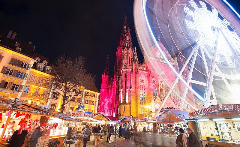 Vue nocturne du marché de Noël de Mulhouse avec la grande roue illuminée et la cathédrale en arrière-plan.