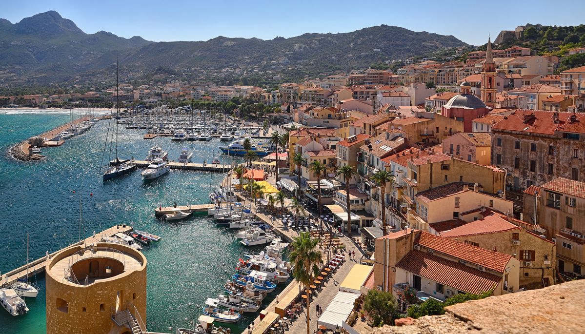 Vue du port de Calvi en Corse, avec ses bateaux amarrés, ses maisons aux toits rouges et les montagnes en arrière-plan par une journée ensoleillée de juillet.