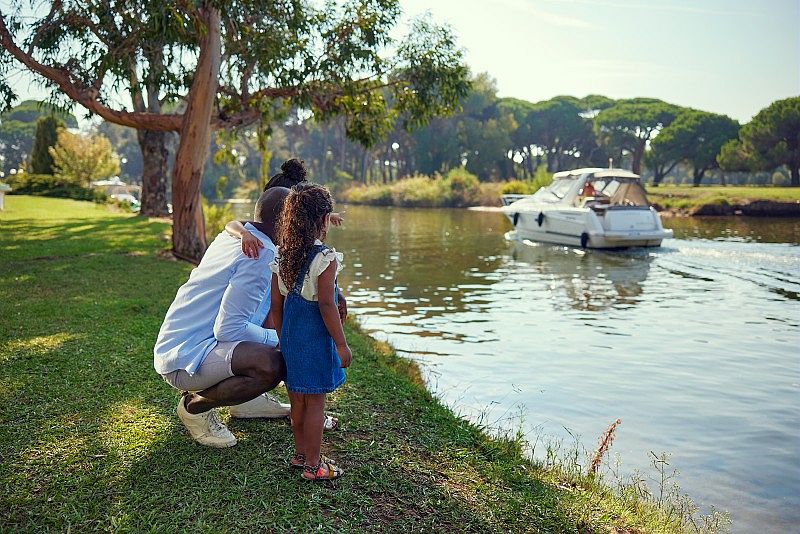 Un homme, une femme et une petite fille regardent un bateau à moteur passer sur un canal. Ils sont accroupis sur l’herbe au bord de l’eau, entourés d’arbres sous un ciel dégagé. La scène est paisible, en pleine nature.