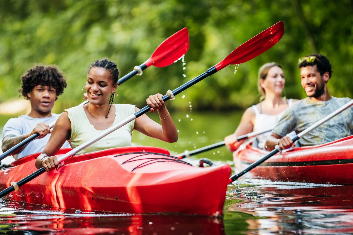 Un groupe d'amis pagaient dans des kayaks rouges sur une rivière entourée de verdure. Tous sourient et profitent d’une activité en plein air sous le soleil.