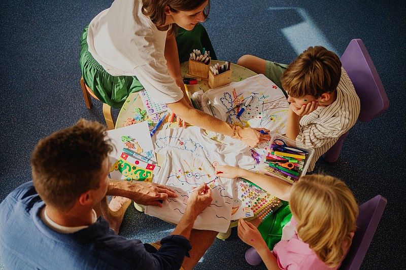 dans le club enfants, deux adultes et deux enfants sont assis autour d’une table ronde et colorient des dessins sur des t-shirts blancs. La table est couverte de feutres, autocollants et papiers colorés, créant une ambiance créative et ludique dans un espace intérieur lumineux