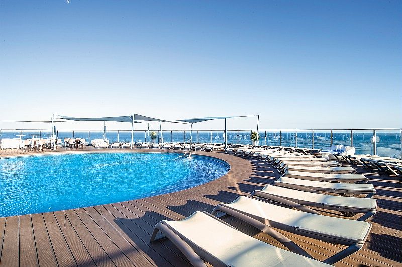 Piscine circulaire sur le rooftop de l’Hôtel El Puerto, entourée de transats et de toiles d’ombrage, avec vue dégagée sur la mer sous un ciel bleu clair.