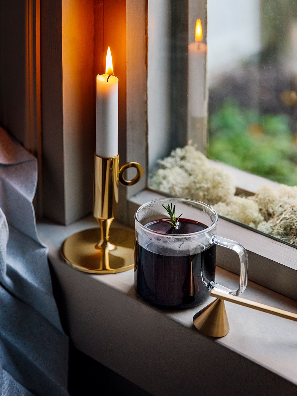 Kuva, jossa ikkunalaudalla kynttilänjalka kynttilöineen, HAYn lasimuki täynnä mustaherukkaglögiä ja ferm LIVINGin messinginvärinen kynttilänsammutin, osana kodin joulusisustusta.