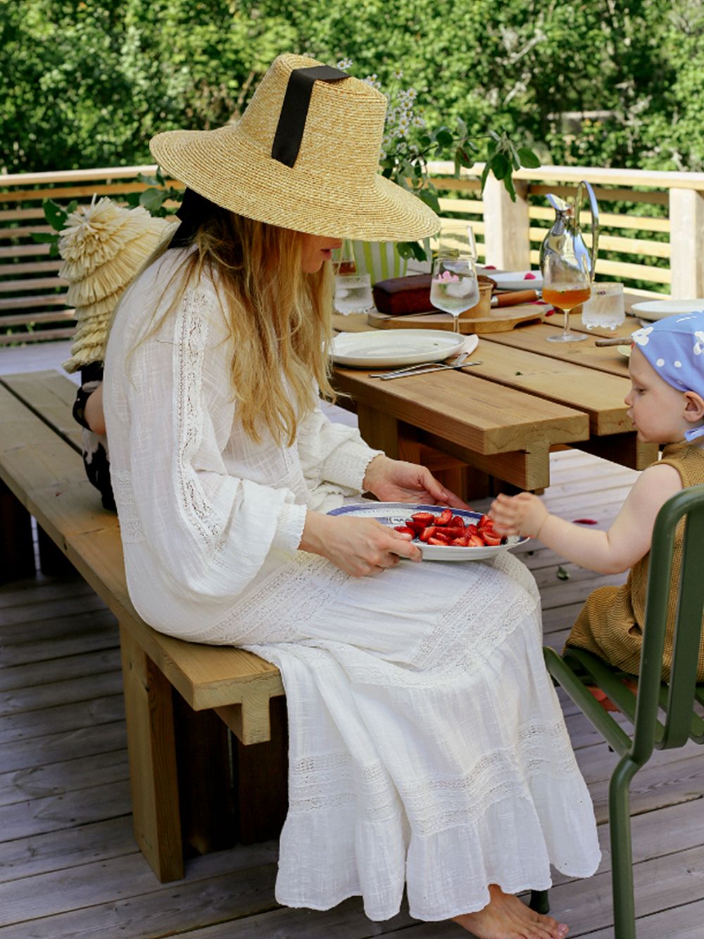 Kuva, jossa Kirsikka Simberg lapsineen kesäisesti katetun mökkipöydän ääressä. Pöytä kuuluu Vaarniin Osa-kokoelmaan.