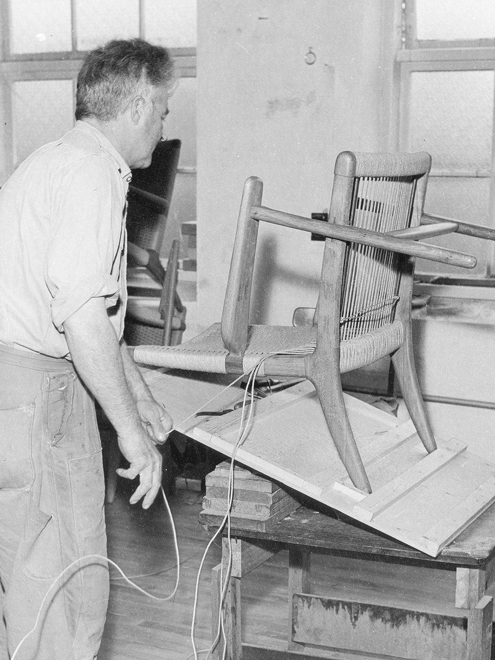 Vanha, mustavalkoinen kuva, jossa artisaani punoo käsin Carl Hansen & Sønin CH25-tuolin istuinosaa.