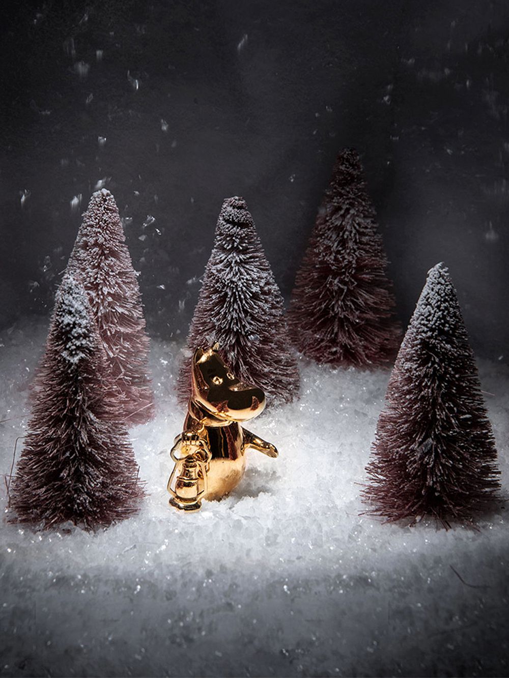 Kullatulla Muumipeikko-figuurilla on kädessään lyhty, ja hän seisoo lumisessa maisemassa kuusipuiden keskellä.