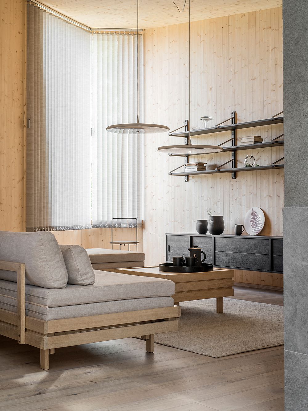 Oleskelutilassa on sohvana Tapio Anttila Collectionin Day&Night-vuodesohva ja sen edessä saman sarjan puinen sohvapöytä. Hämärässä huoneessa on puunväriset seinät ja ikkuna, jota peittävät sälekaihtimet. 