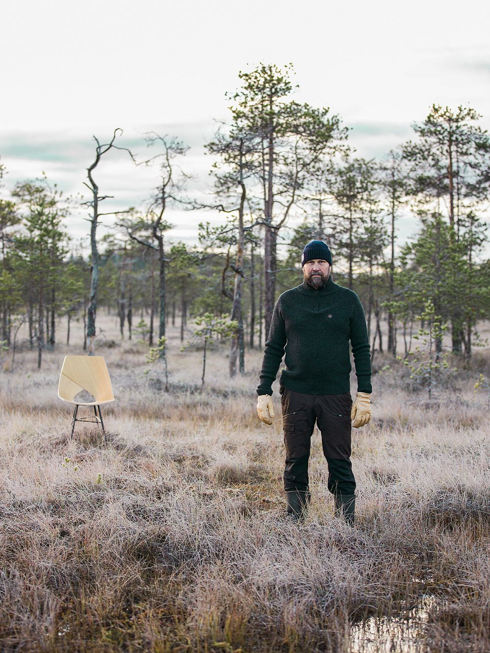 Muotoilija Tapio Anttila seisoo metsässä mustat vaatteet ja pipo yllään. Taustalla Limi-tuoli.