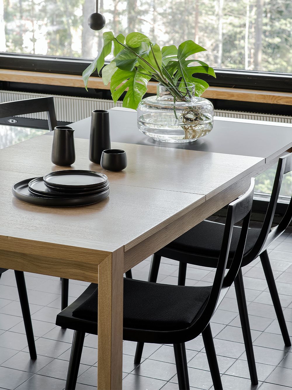 Tapio Anttila Collectionin Jat-ko-tuokapöydän ympärillä Viiva-tuolit. Pöydälle on nostettu lasimaljakko, jossa vihreitä peikonlehden oksia, sekä mustia keraamisia astioita.