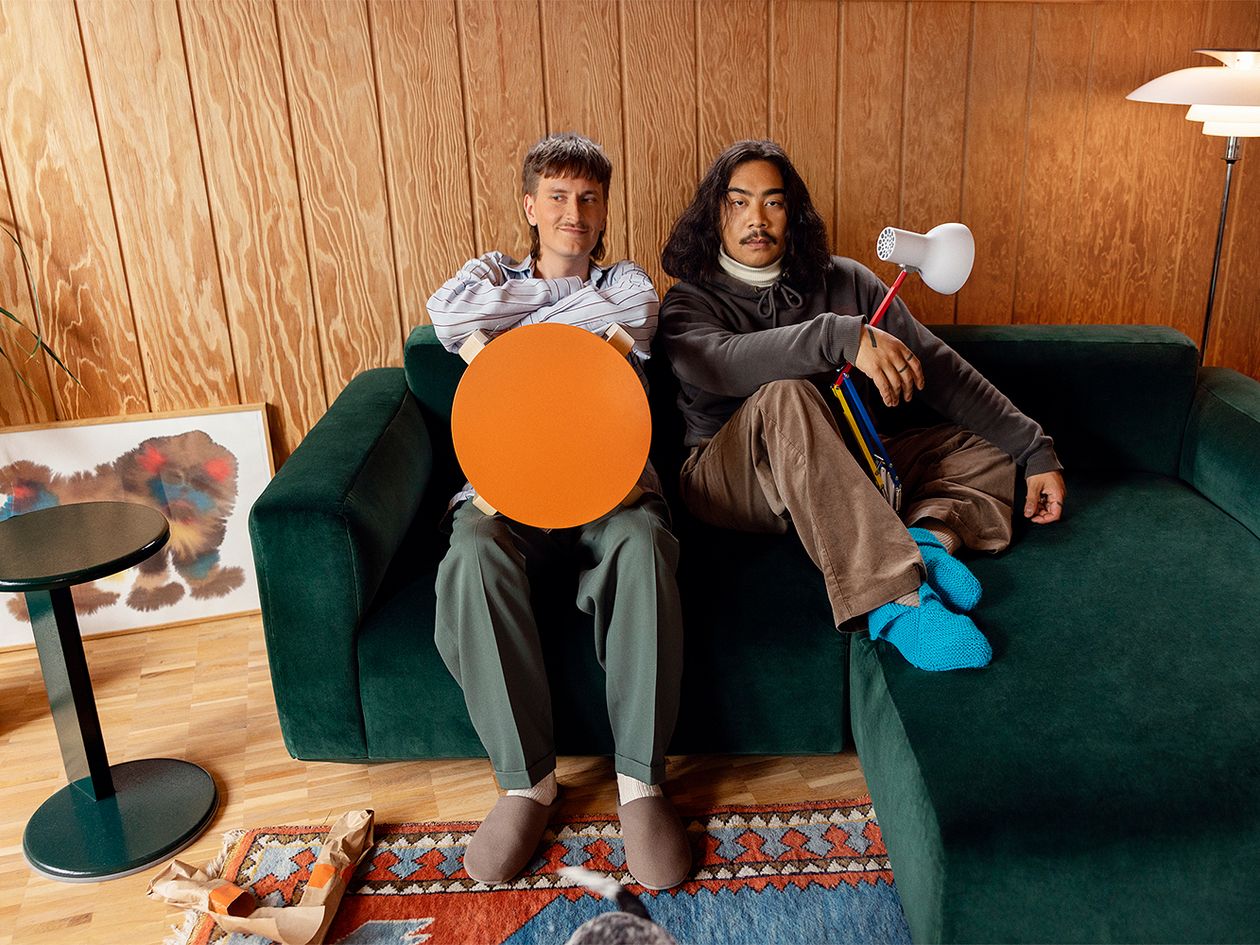 Kuva, jossa kaksi henkilöä istuu vihreällä sohvalla. Toinen pitää sylissään Artekin oranssia jakkaraa, toinen Anglepoisen Type 75 Mini -pöytävalaisinta.