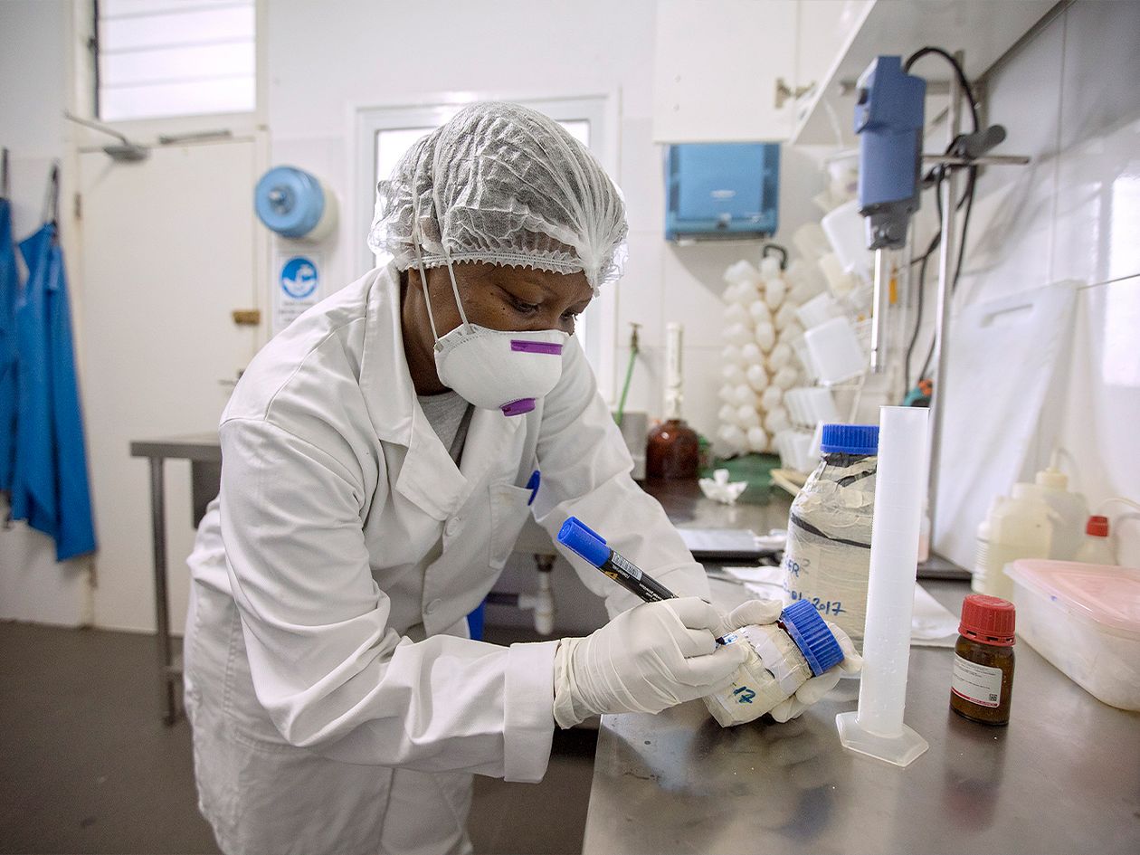Nainen työskentelee laboratoriossa yllään valkoinen takki ja päässään suojapäähine.