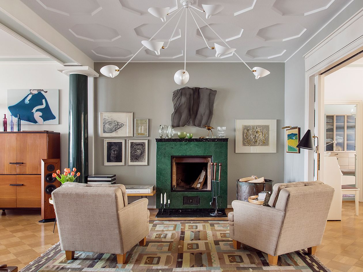 An image taken in Aija Staffan's livingroom. There is a green fireplace, a vibrant rug, two lounge chairs, and many pieces of art on the walls.