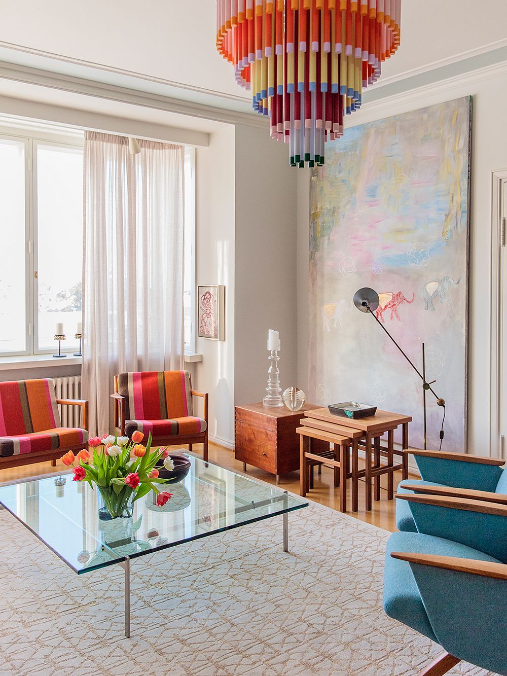 An image of a living room: a glass coffee table, a colorful ceiling lamp, colorful vintage lounge chairs and a large painting on the wall.