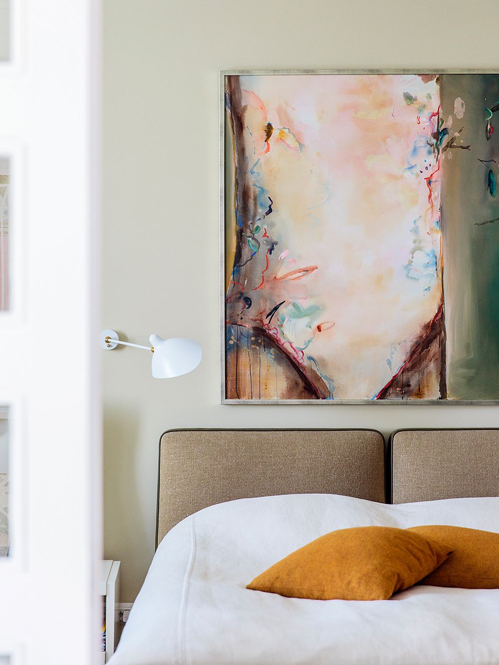 An image of a bedroom: a bed, some throw pillows, a wall lamp, a painting on the wall.