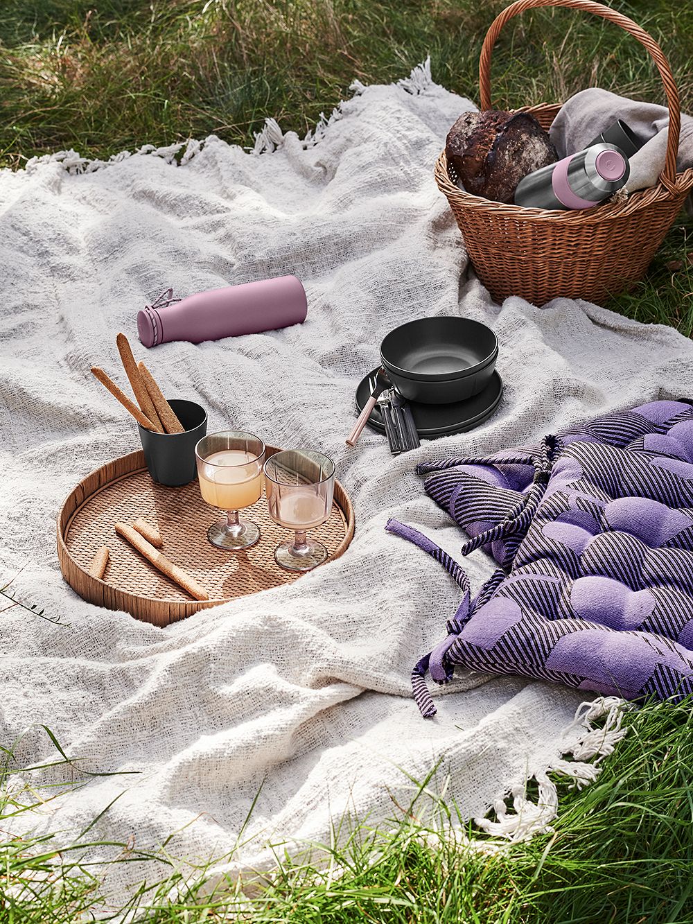 Ruohikolle aseteltu piknikviltti, jonka päälle on asetettu violetti Grand Cru -termospullo, pyöreä tarjotin, ruokia, istuintyyny sekä rottinkinen piknikkori