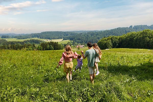 Une famille de quatre personnes marche dans une prairie verdoyante en été. Les parents tiennent leurs enfants par la main, tournés de dos, en direction d’un paysage vallonné avec des forêts et des champs à l’horizon sous un ciel bleu légèrement nuageux.