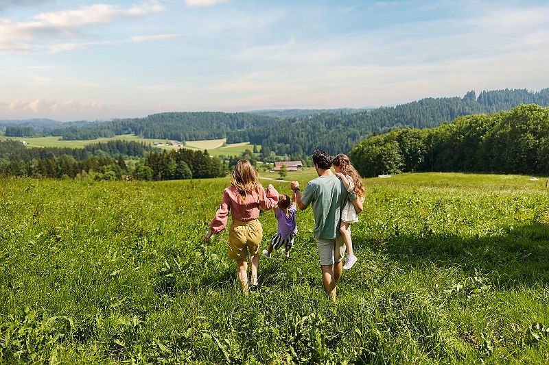 Une famille de quatre personnes marche dans une prairie verdoyante en été. Les parents tiennent leurs enfants par la main, tournés de dos, en direction d’un paysage vallonné avec des forêts et des champs à l’horizon sous un ciel bleu légèrement nuageux.