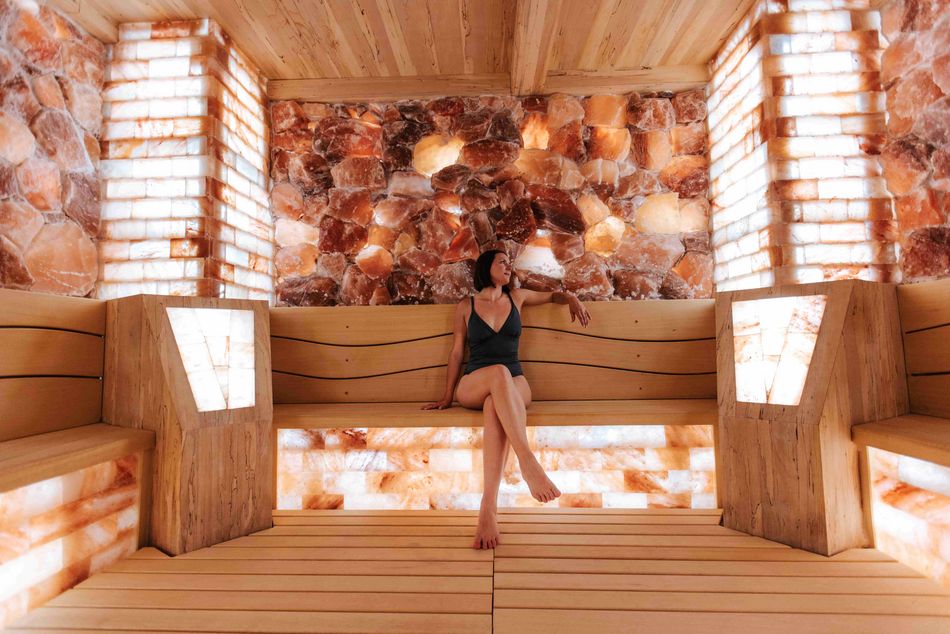 Une femme en maillot de bain assise sur un banc en bois dans une salle de sel, entourée de murs en briques de sel illuminées. L'ambiance est apaisante et relaxante.