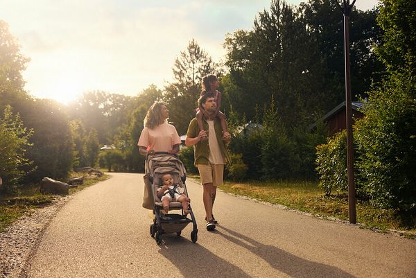 Une famille se promène sur un chemin verdoyant. La mère pousse une poussette avec son bébé dedans et le père marche à côté d'elle avec sa fille sur ses épaules.