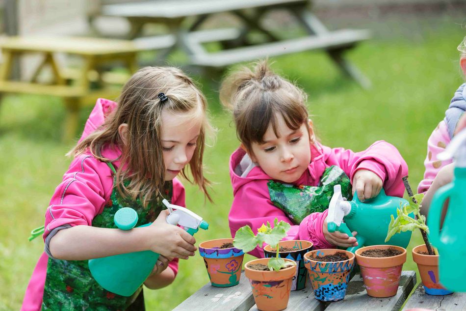 Deux jeunes filles portant des tabliers verts et des vestes roses arrosent de petites plantes dans des pots décorés avec des pulvérisateurs verts. Elles sont concentrées sur leurs activités de jardinage en plein air.