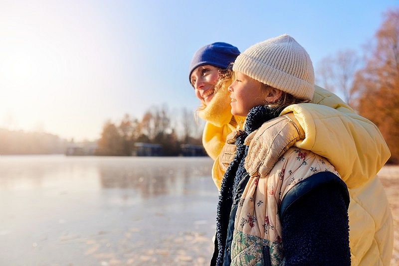 Une mère en bonnet bleu enlace son enfant en tenue d’hiver au bord d’un lac gelé, sous une lumière douce de fin de journée.