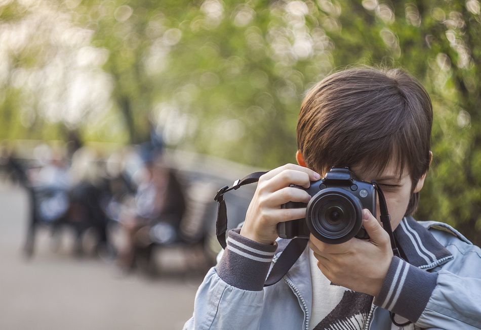 Un jeune garçon tenant un appareil photo et prenant une photo dans un parc, avec un fond flou de personnes assises sur des bancs et de verdure.