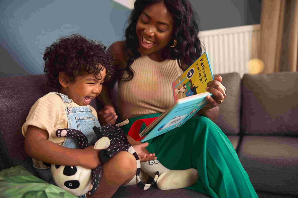 Une femme souriante lit un livre illustré à un jeune enfant assis sur un canapé, qui rit en tenant une peluche. Moment de partage et de complicité lors d'une lecture.