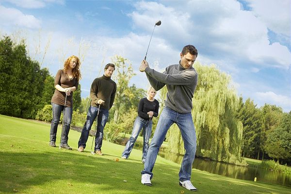 Un groupe d'amis pratique le golf sur un terrain verdoyant, sous un ciel bleu. L'un d'eux s'apprête à frapper la balle tandis que les autres observent, créant une ambiance conviviale et détendue.