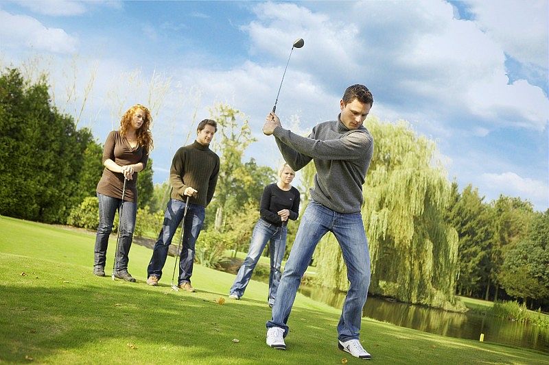 Un groupe d'amis pratique le golf sur un terrain verdoyant, sous un ciel bleu. L'un d'eux s'apprête à frapper la balle tandis que les autres observent, créant une ambiance conviviale et détendue.