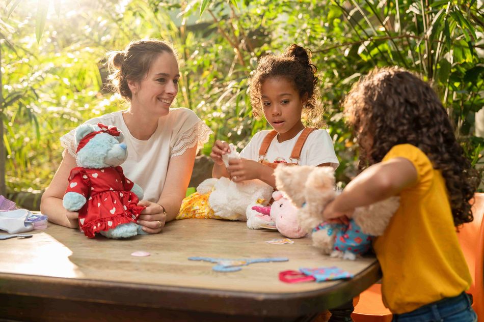 Une femme souriante assise à une table avec deux enfants jouant avec des peluches. Les enfants habillent leurs jouets, entourés de vêtements et accessoires colorés, dans un cadre verdoyant et ensoleillé.