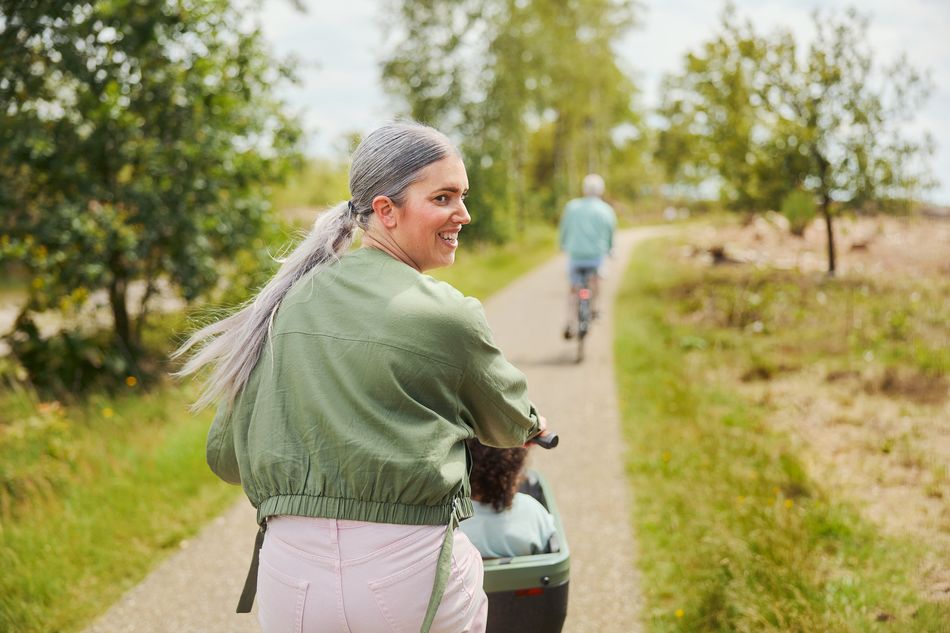 Une femme à vélo avec un enfant dans un siège avant, sur un chemin de campagne, souriante et regardant en arrière.