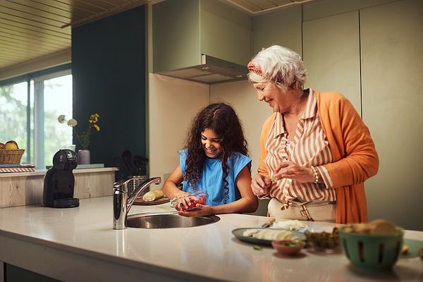 Une femme âgée et une jeune fille sourient en préparant des fruits ensemble dans une cuisine moderne. La jeune fille lave des fraises sous le robinet pendant que la femme l'observe avec tendresse.