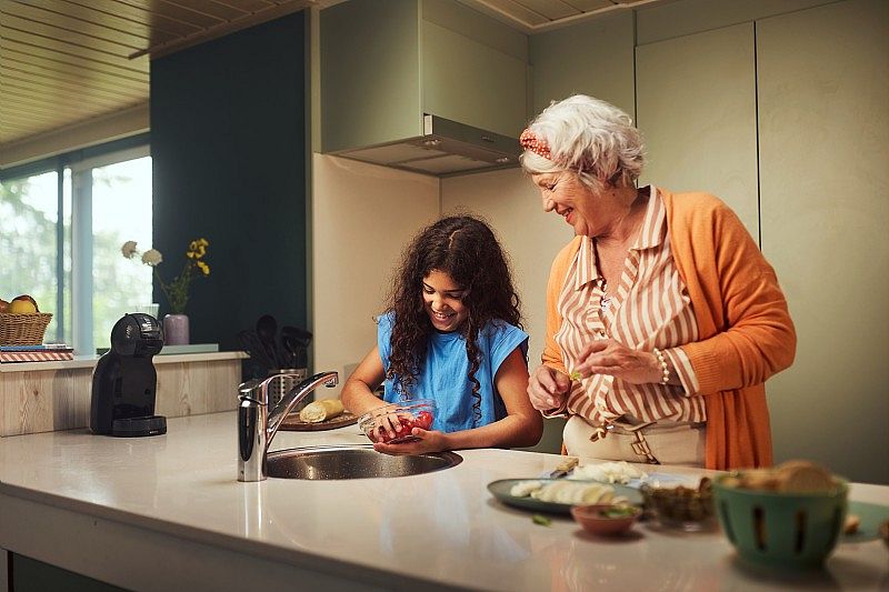Une femme âgée et une jeune fille sourient en préparant des fruits ensemble dans une cuisine moderne. La jeune fille lave des fraises sous le robinet pendant que la femme l'observe avec tendresse.