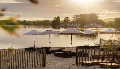 Plage aménagée au bord d’un lac paisible au coucher du soleil, avec des transats en bois et des parasols blancs alignés, face à l’eau calme. À l’horizon, une île boisée et quelques maisons sont baignées par la lumière dorée du soir.