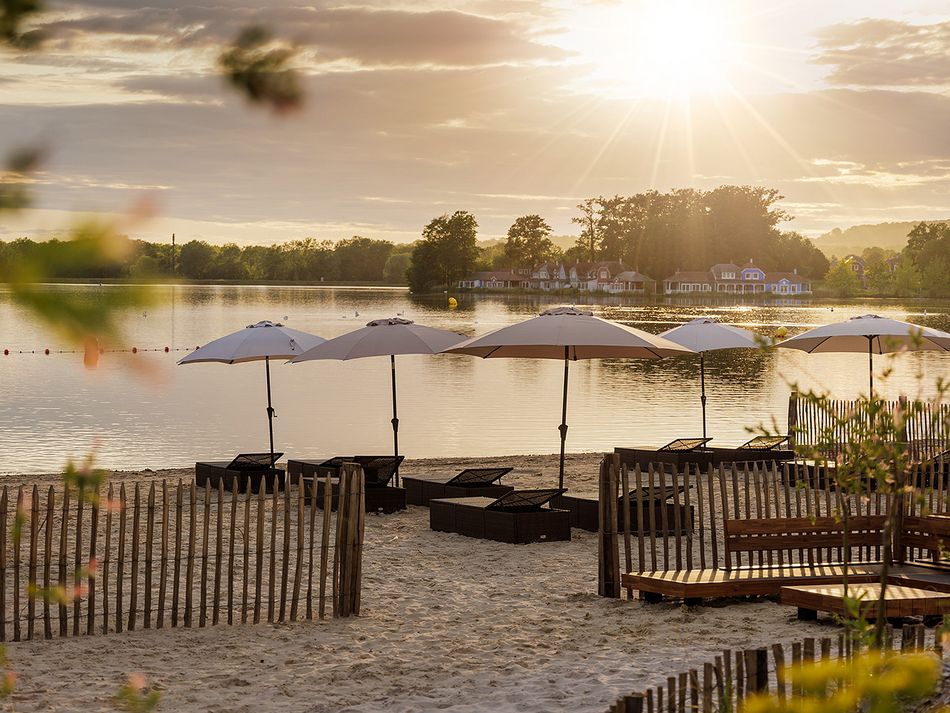 Plage aménagée au bord d’un lac paisible au coucher du soleil, avec des transats en bois et des parasols blancs alignés, face à l’eau calme. À l’horizon, une île boisée et quelques maisons sont baignées par la lumière dorée du soir.