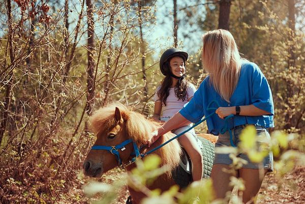 Une enfant souriante monte un poney dans une forêt ensoleillée, accompagnée d'une adulte qui tient les rênes. L’ambiance est chaleureuse, avec des rayons de soleil filtrant à travers les arbres et des feuillages printaniers autour.