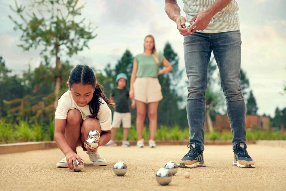 Une jeune fille ramasse des boules de pétanque sur un terrain, pendant qu’un adulte se prépare à lancer. En arrière-plan, une femme et un enfant regardent la partie.