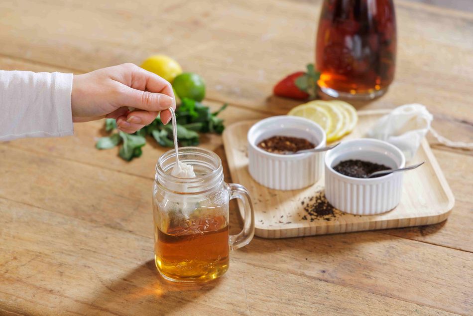 Une main plonge un sachet de thé dans un verre en verre rempli d'eau chaude. En arrière-plan, une planche avec des bols de thé en vrac, des tranches de citron, des herbes et une carafe de thé glacé.